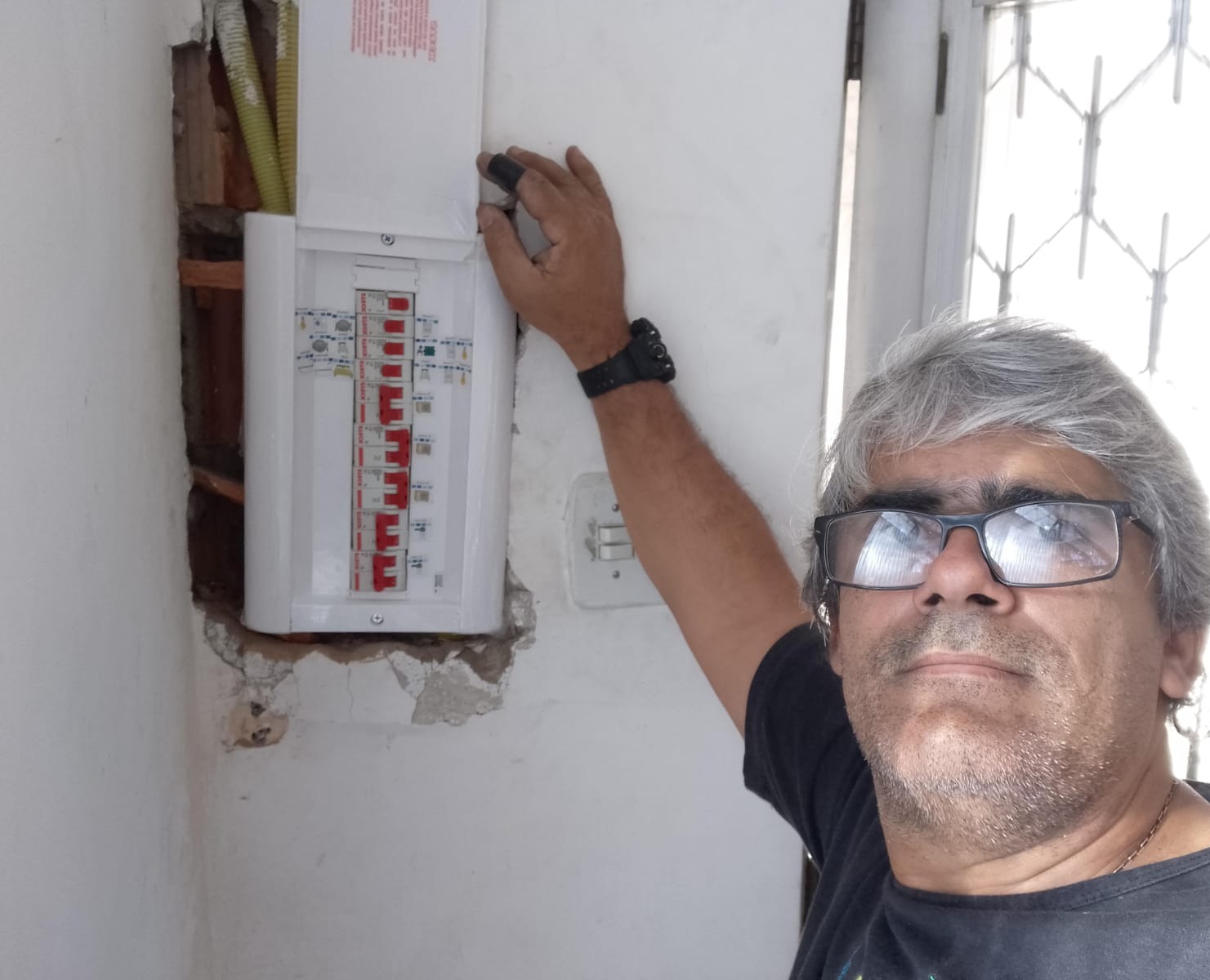 homem de cabelos grisalhos e óculos tirando selfie ao lado de um quadro de disjuntores aberto na parede